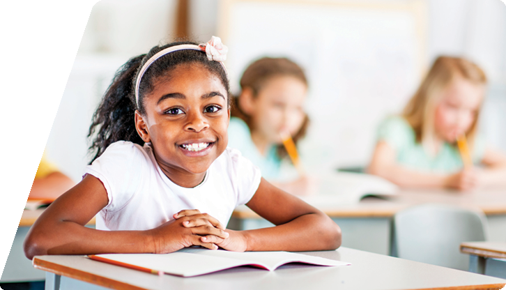 Foto de menina de cerca de sete anos, sentada em uma carteira escolar, com os ombros apoiados sobre um livro aberto. Ela sorri e olha para a câmera. A menina tem cabelos crespos presos e usa tiara e blusa branca. Ao fundo, duas colegas de classe escrevem.