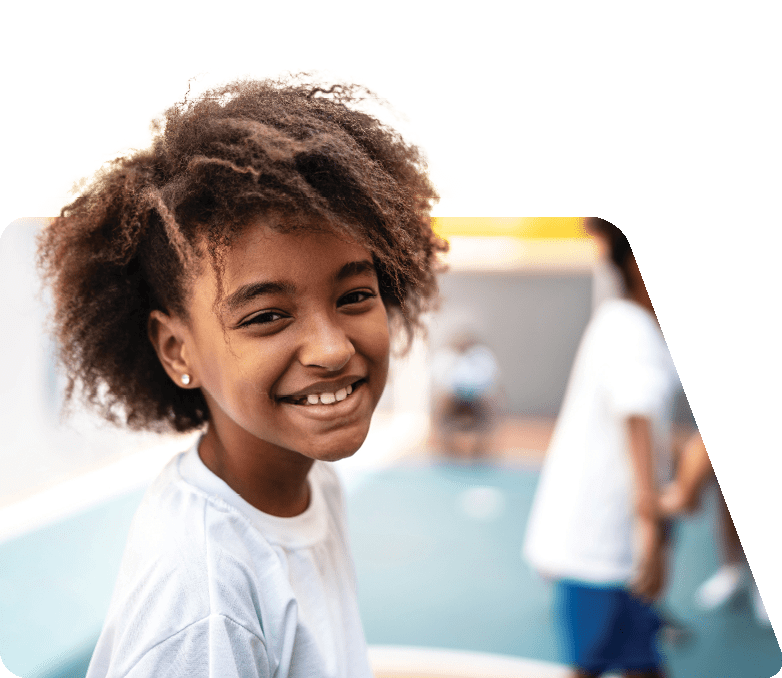 Foto de menina negra, de cerca de 10 anos, com cabelos crespos, acima da altura do ombro. Ela veste camiseta branca, sorri e olha para a câmera.